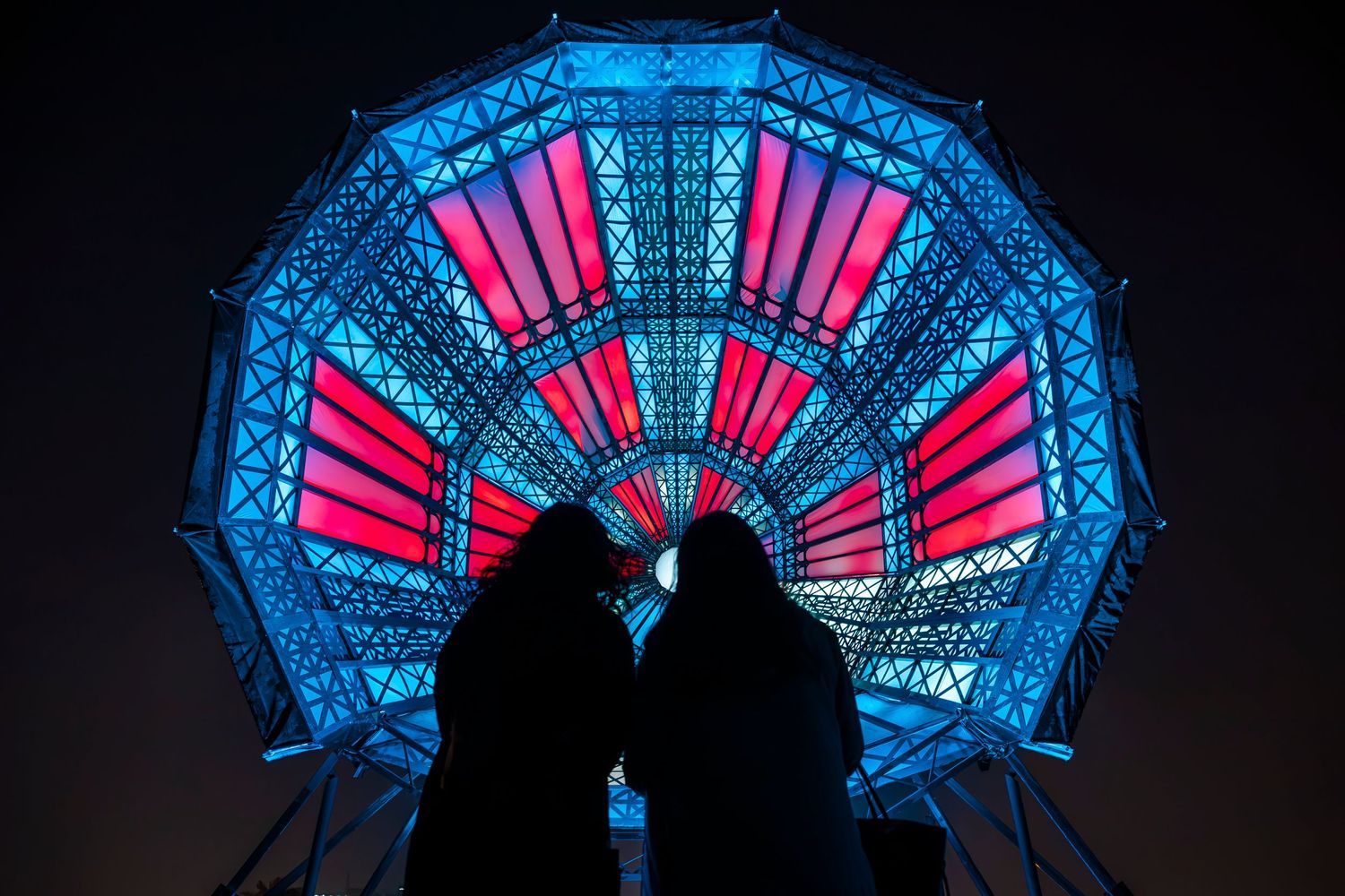 Silhouettes stand before a geometric dome illuminated in vibrant blue and pink lighting patterns.