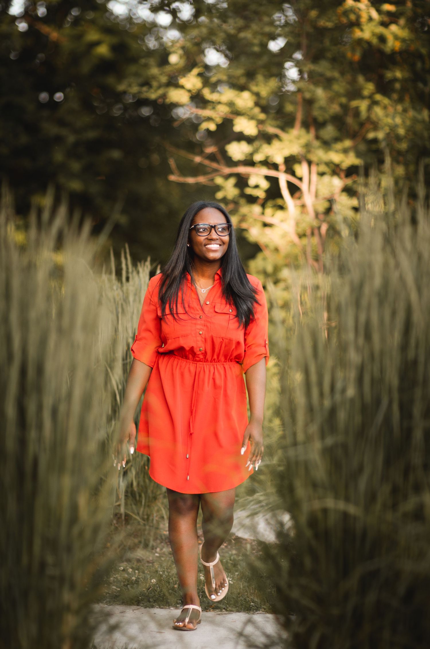 Woman in bright coral dress walking along path through tall grass at golden hour.