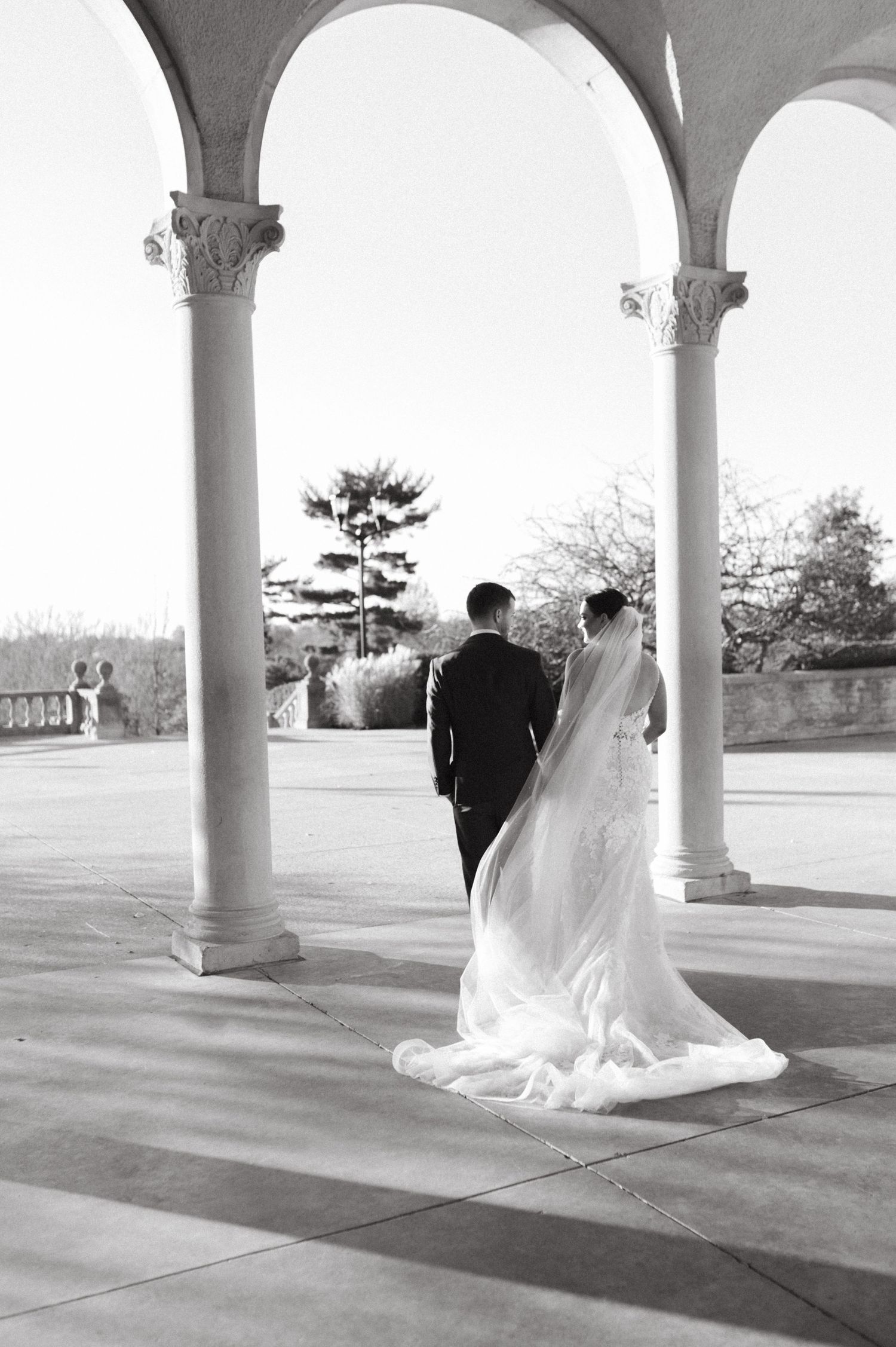 Black and white photo of couple under elegant arched columns with flowing wedding veil.
