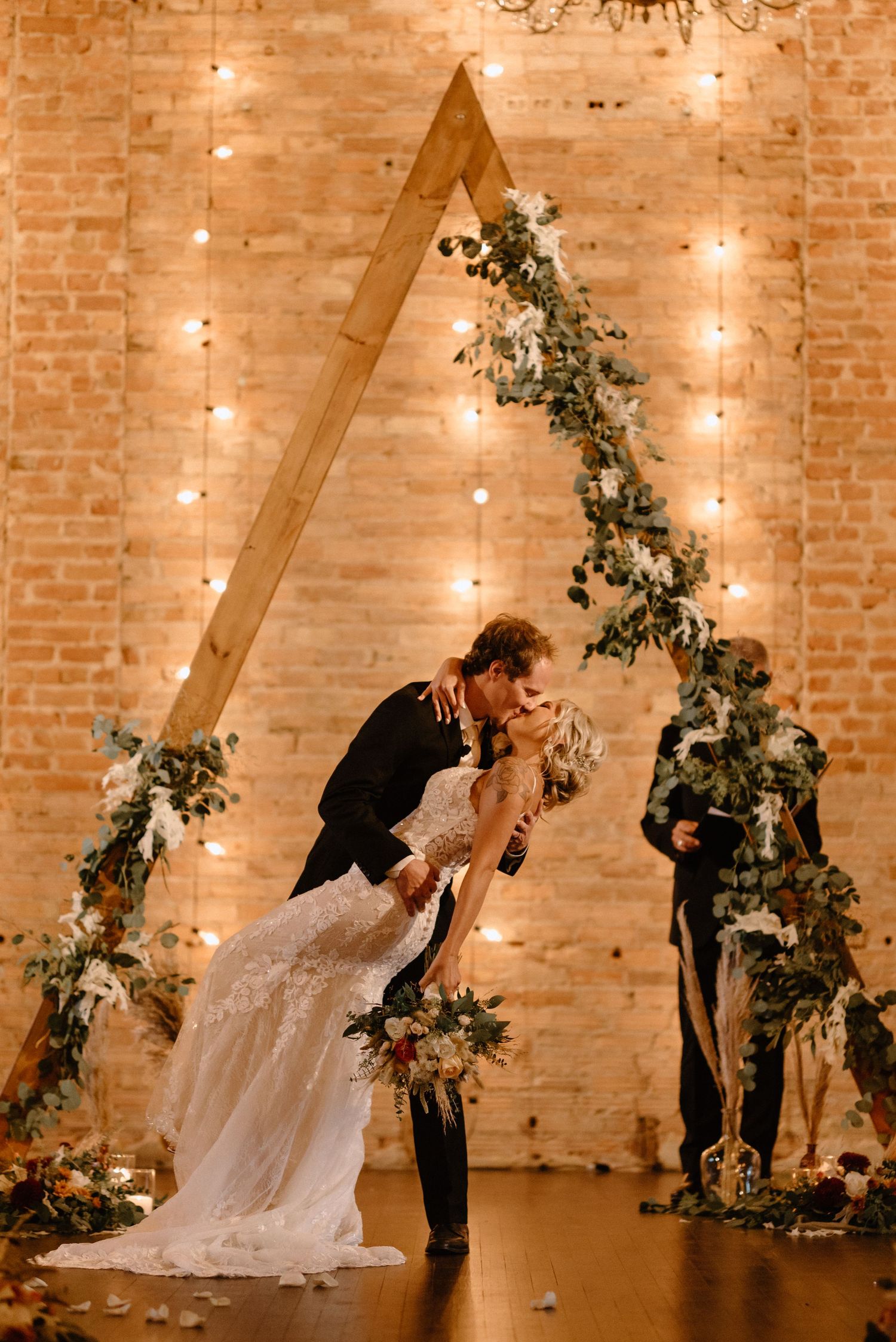 Romantic wedding couple dips beneath triangular floral arch with twinkling lights against exposed brick wall.