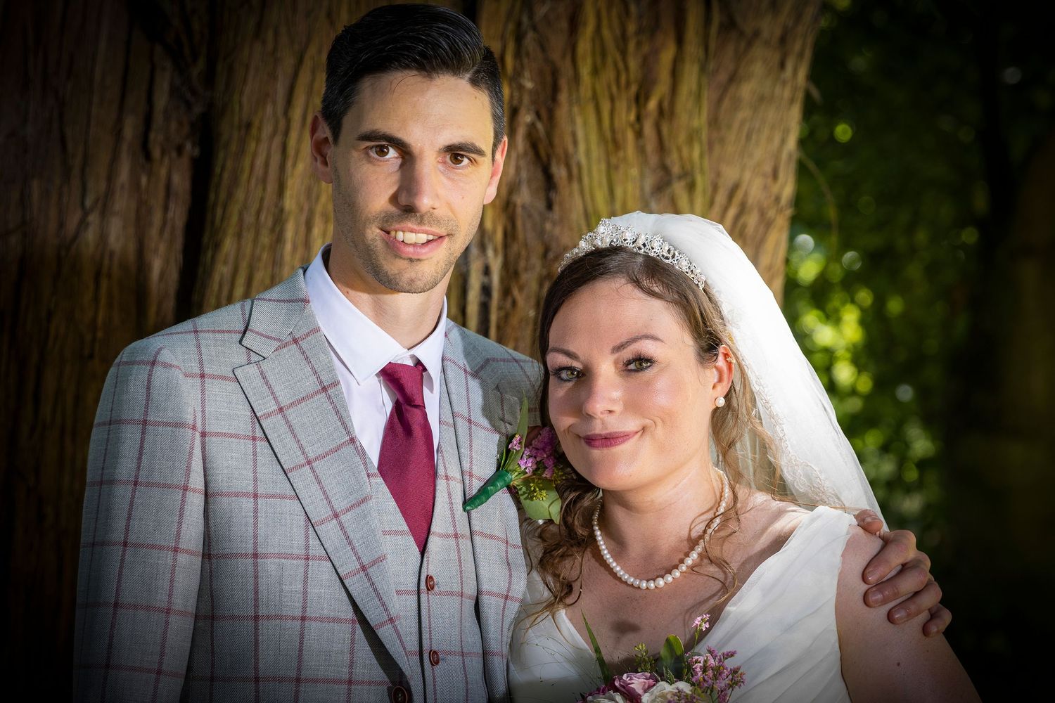 A romantic outdoor wedding portrait featuring the newlyweds standing by a large tree trunk.