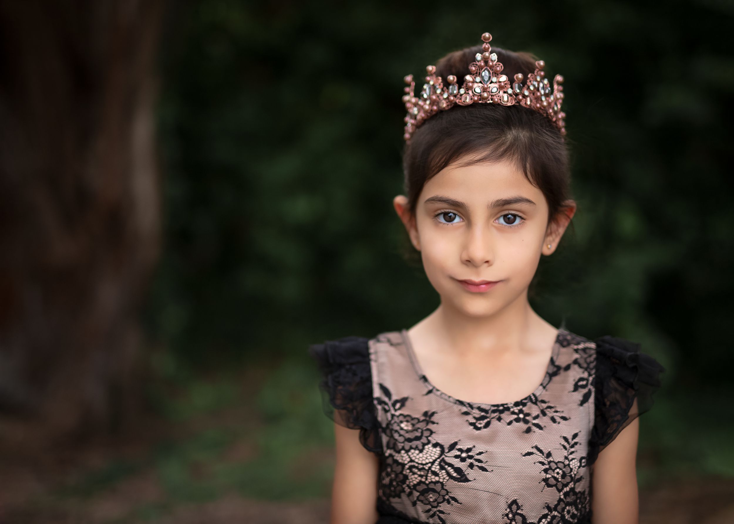 Young girl wearing a crown and black lace gown standing off centre with a dark background of greenery.