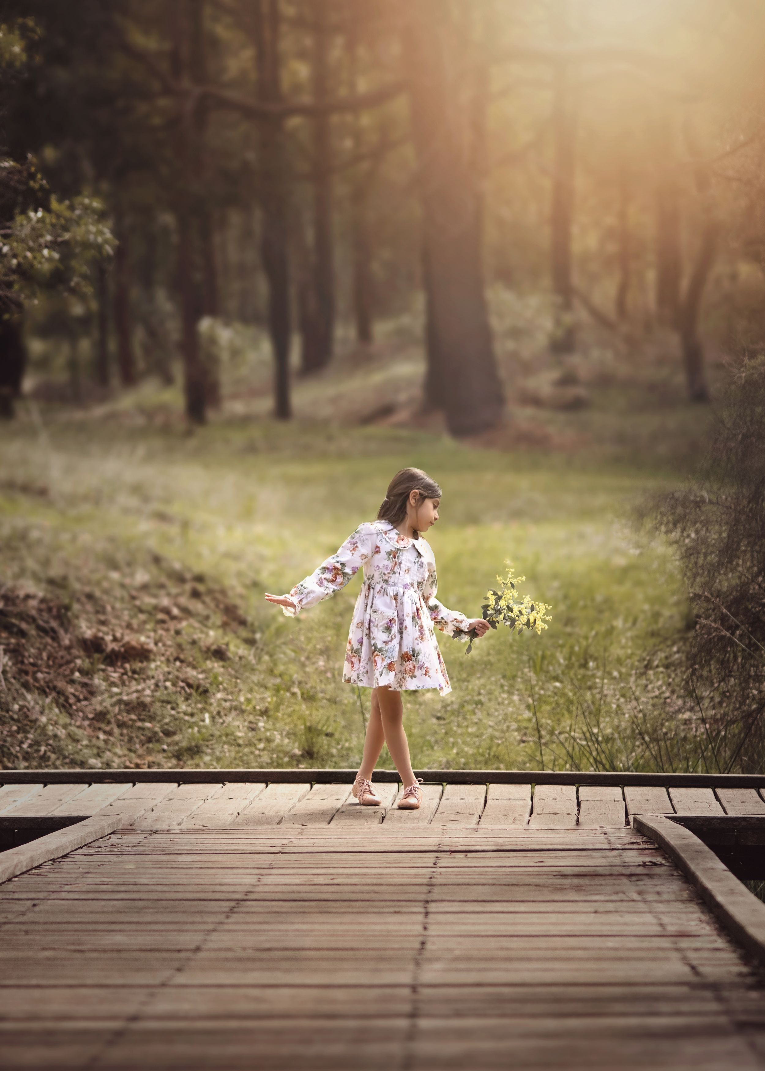 Young girl in vintage dress holding bunch of wattle dancing on a wooden deck in front of a forest at Jarrahdale.