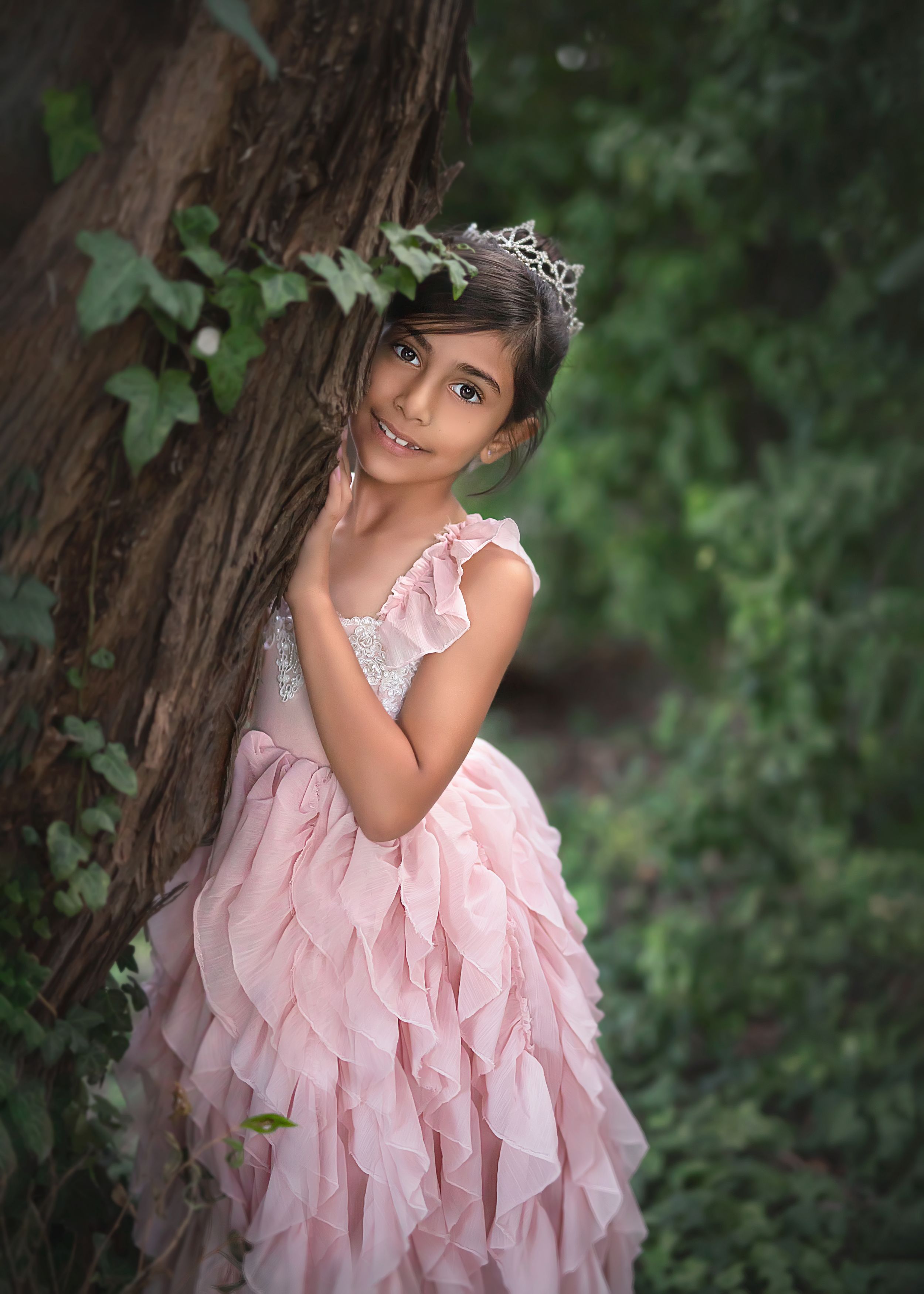 Girl in pink ruffle gown and crown resting against large tree trunk surrounded by lush green ivy. Dream Dress Session.