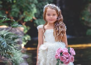 Small girl in a white ruffle gown and beaded headpiece, holding flowers while standing in front of waterfall.