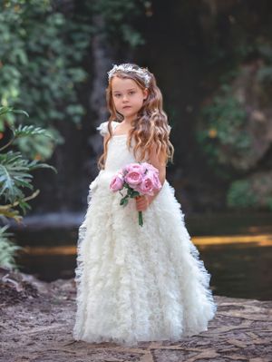 Girl with cascading long brunette hair, in a white gown, holding a bouquet of flowers, standing in front of a waterfall.