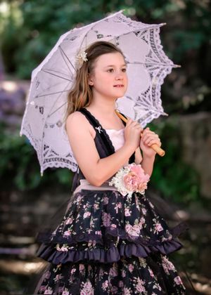Preteen girl in black and pink floral tiered gown holding an open lace parasol over shoulder while looking into distance