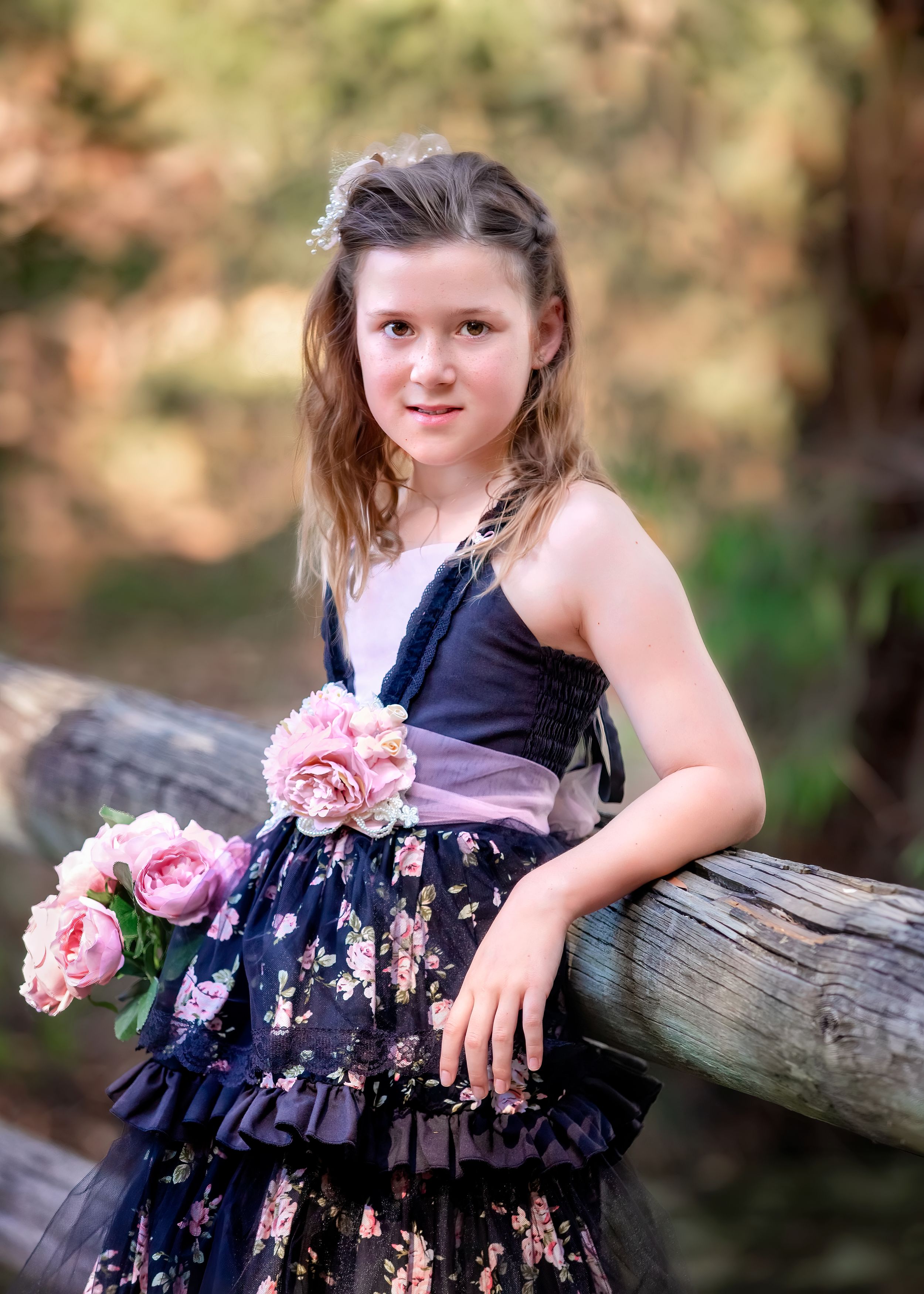 Girl in black and pink floral gown holding flowers leaning back against log railing at John Oldham Park Perth.