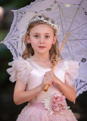 Girl child wearing a beaded hair wreath and floral sash over a pink gown holding an open lace parasol.