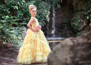 Young girl in yellow ruffled gown standing side on in front of waterfall with hand on hip and smiling at viewer