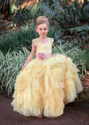 Blonde girl with hair up in bun, standing in front of greenery. Wearing a yellow ruffled gown with pink floral sash.