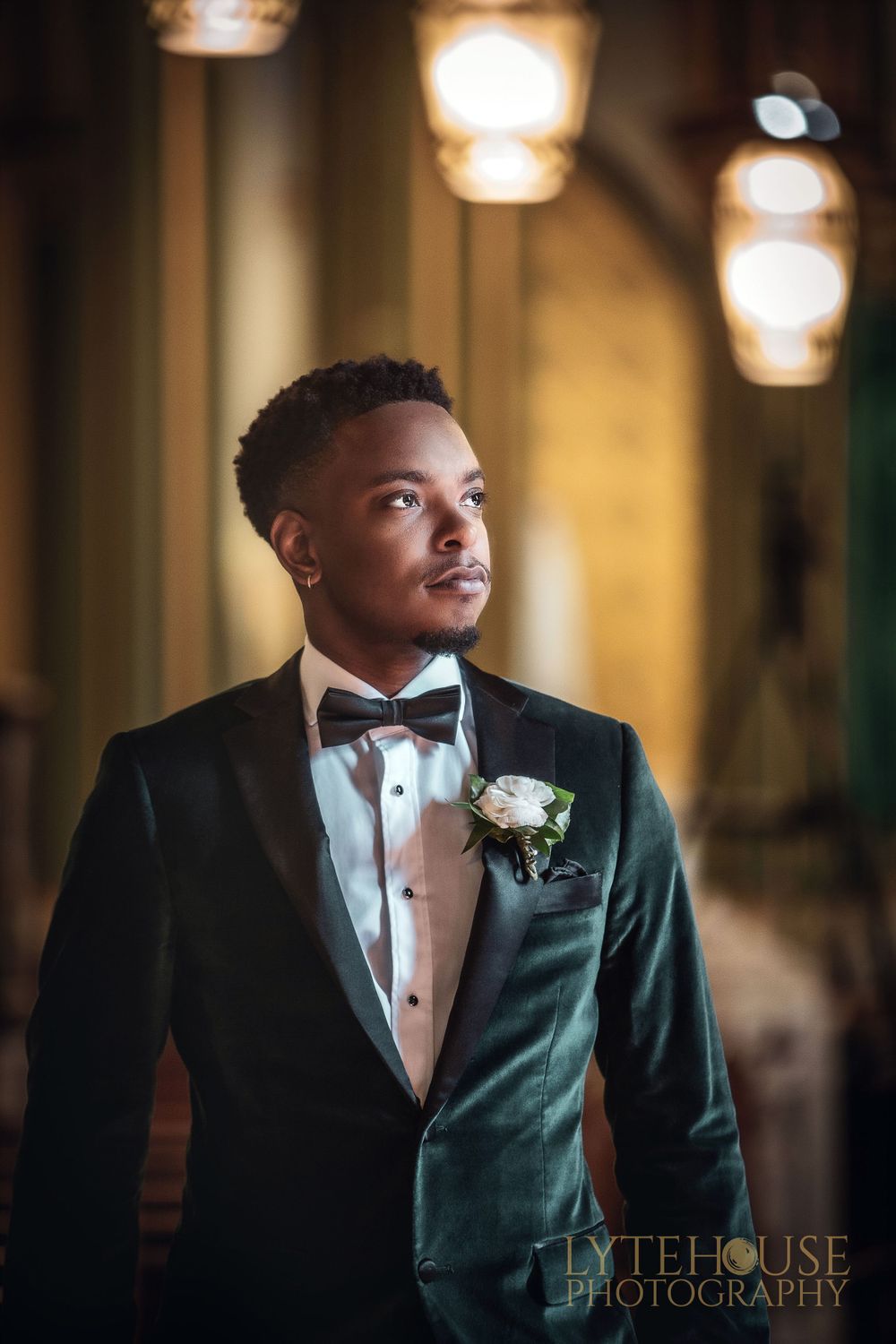 An elegant wedding photo of a groom in a dark velvet tuxedo with bowtie and boutonniere in warm lighting against a blurred hall.