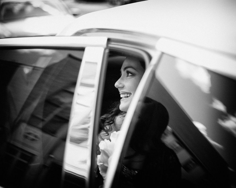 A candid, monochrome photo captures the radiant smile of a bride as she sits inside a car