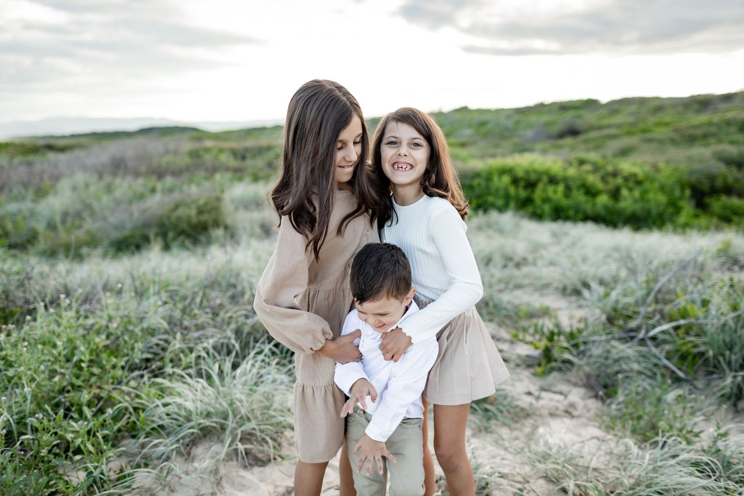 Three people share a candid moment while posing on coastal dunes in neutral-colored clothing.