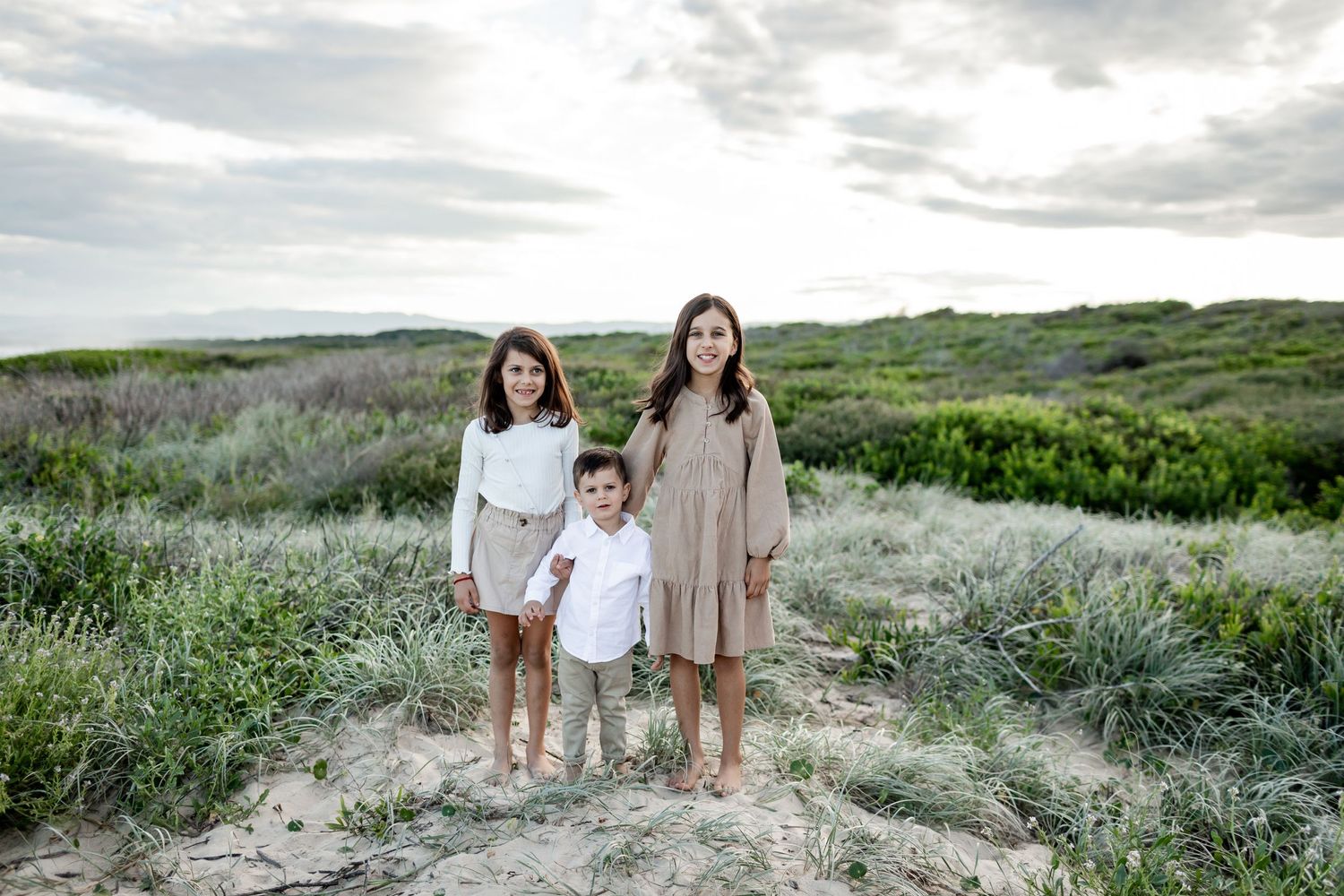 Three people in neutral-toned outfits pose barefoot on coastal dunes with greenery in the background.
