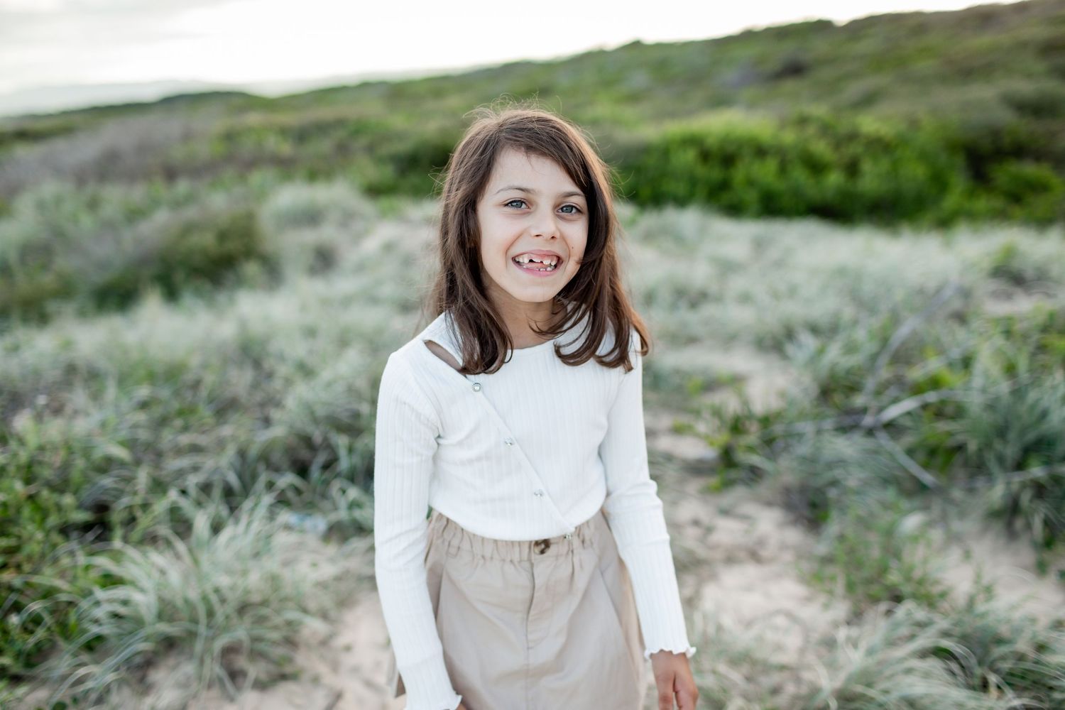 Child wearing white top and khaki pants stands in coastal grassland setting.
