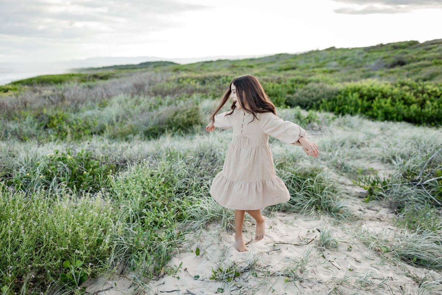 Young child in a cream dress twirls joyfully in a coastal field of grass and sand.