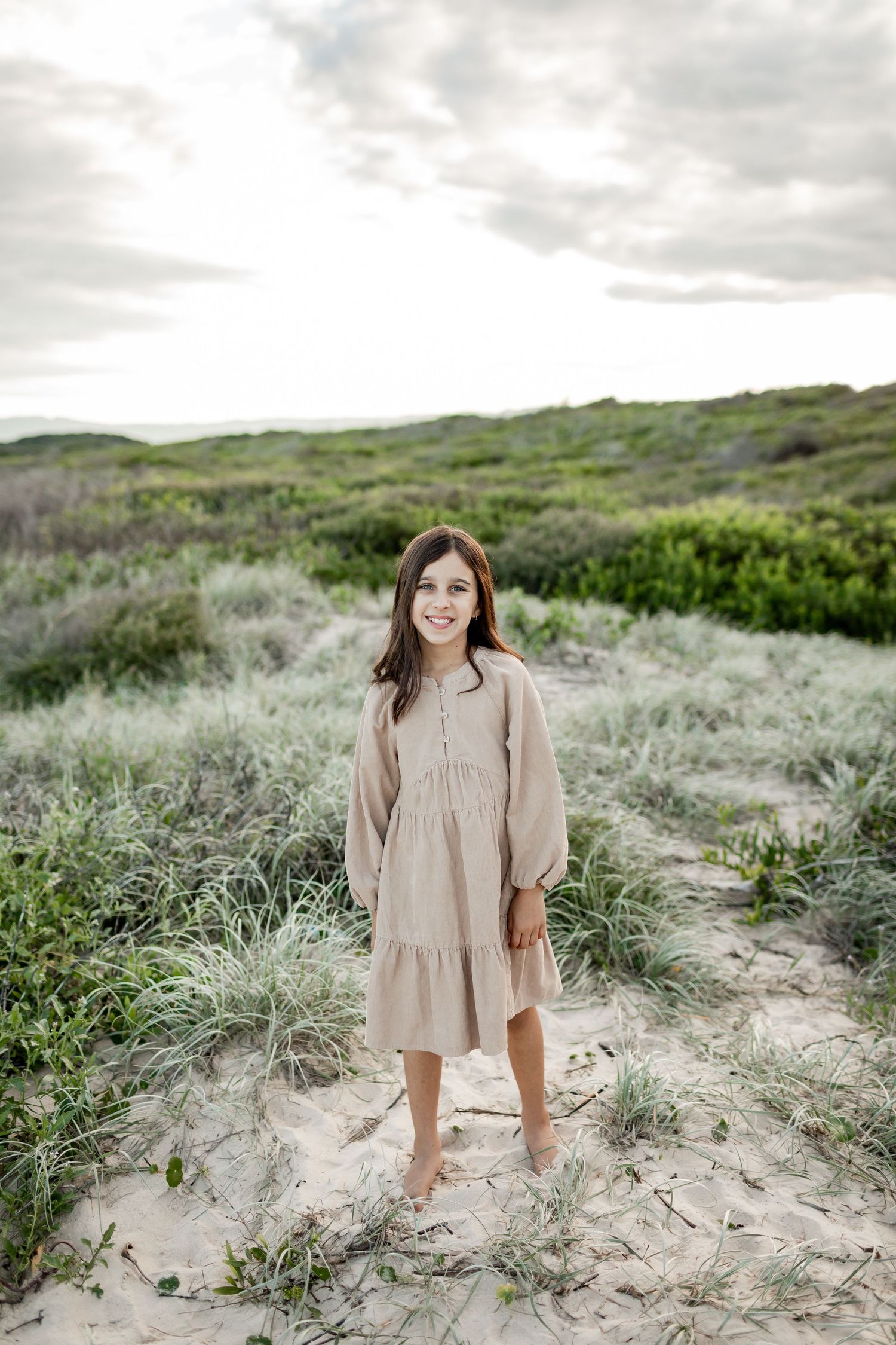 A person in a beige dress stands barefoot on sandy dunes against a cloudy coastal landscape.