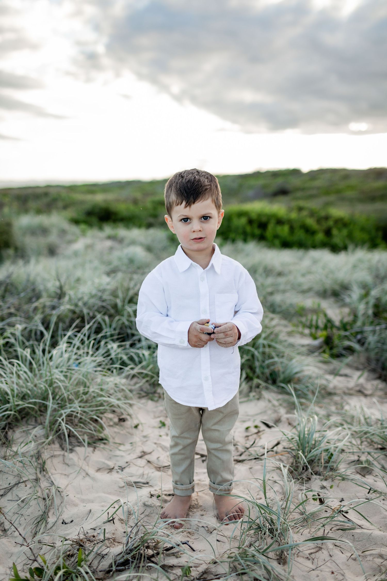 Small child in white shirt and khaki pants stands barefoot on sandy ground.