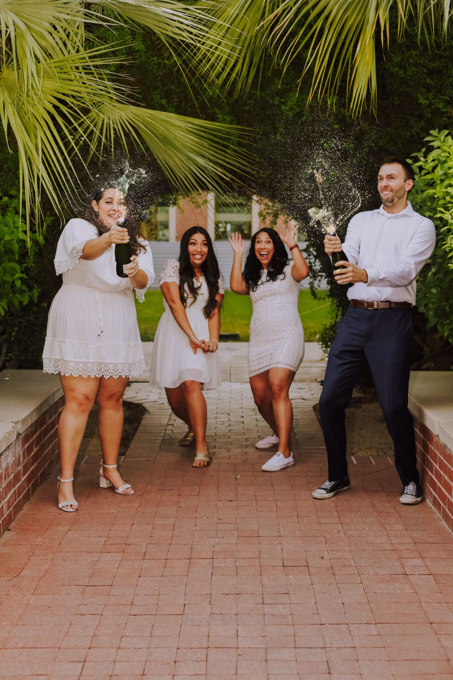 A celebratory group spraying champagne outdoors while wearing white dresses and formal attire.