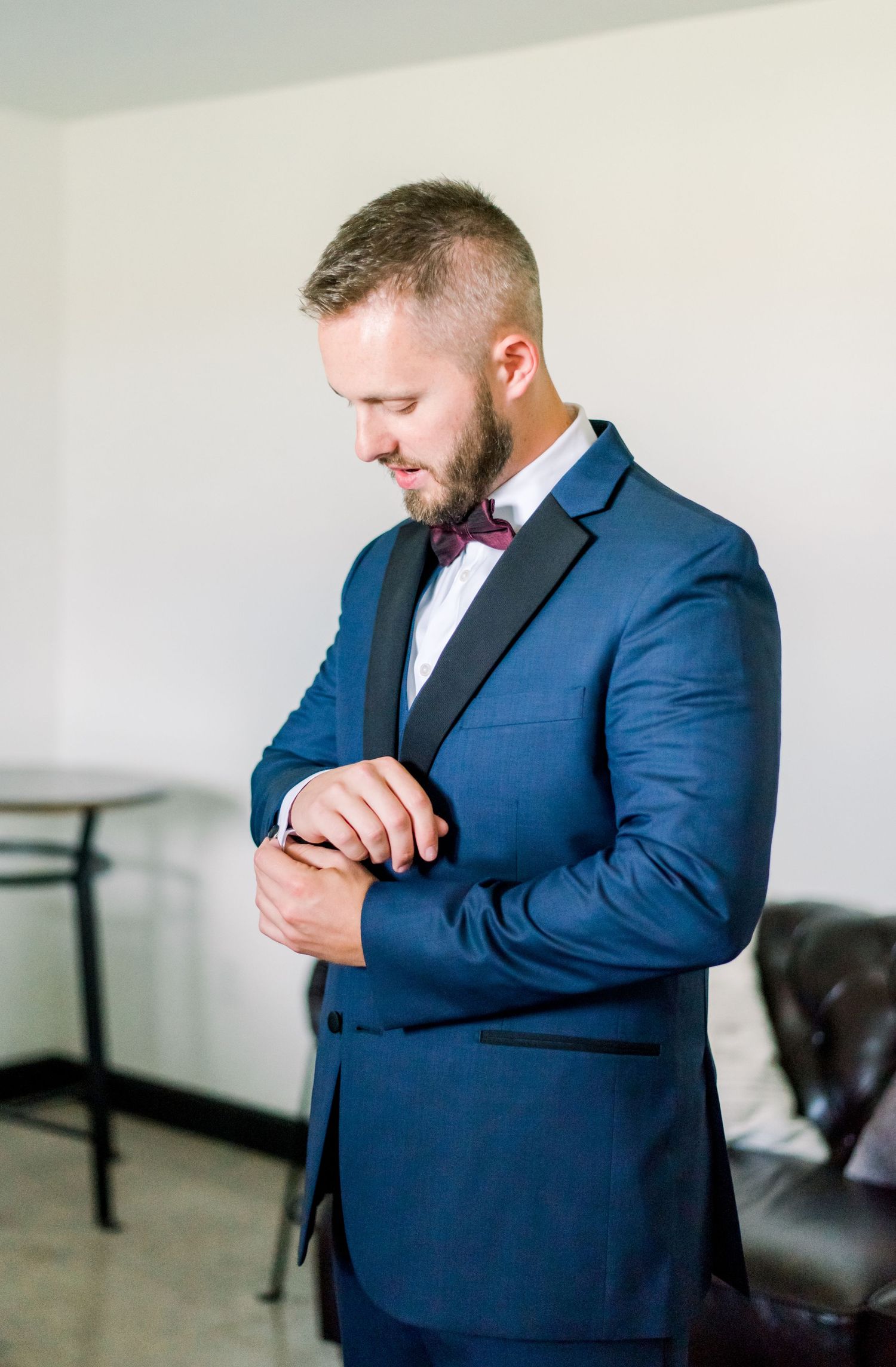 Groom in navy blue suit and burgundy bow tie adjusting cufflinks.