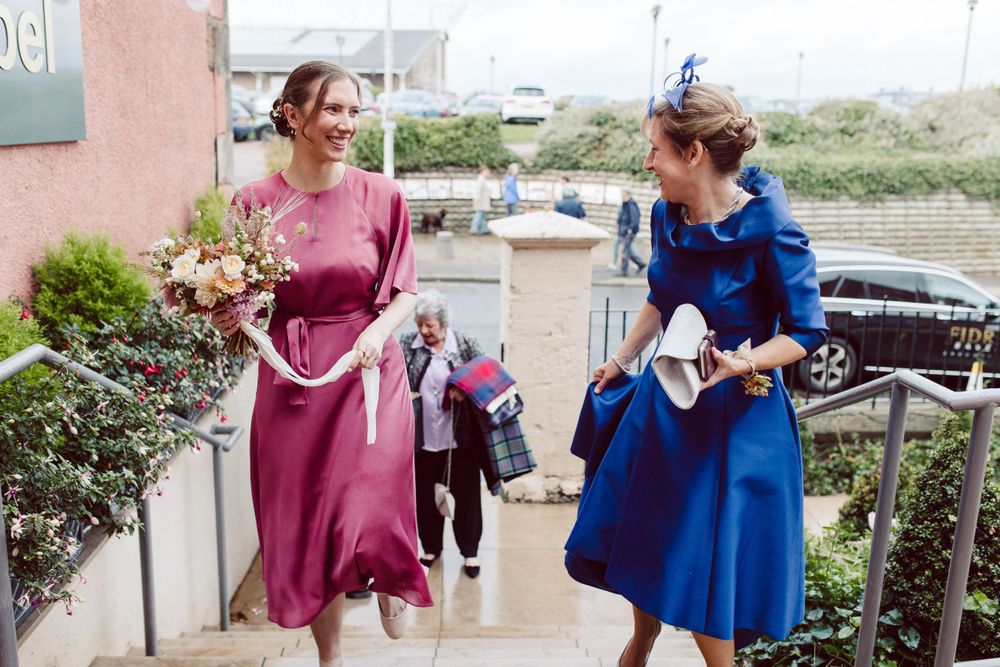Two attendees in elegant dresses, one burgundy and one royal blue, hold bouquets while walking outdoors.