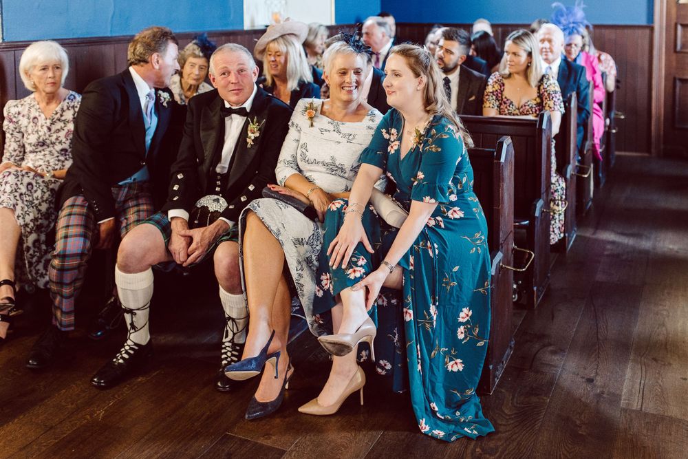 Wedding guests in formal attire seated on wooden benches during a ceremony.