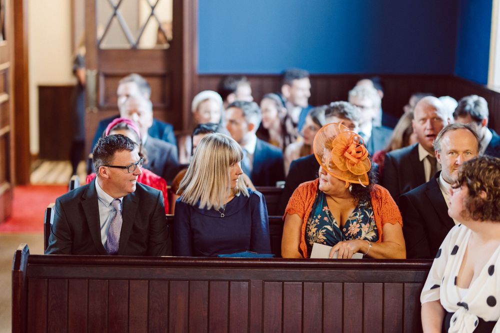 Wedding guests seated in wooden pews with bright blue walls in the background.