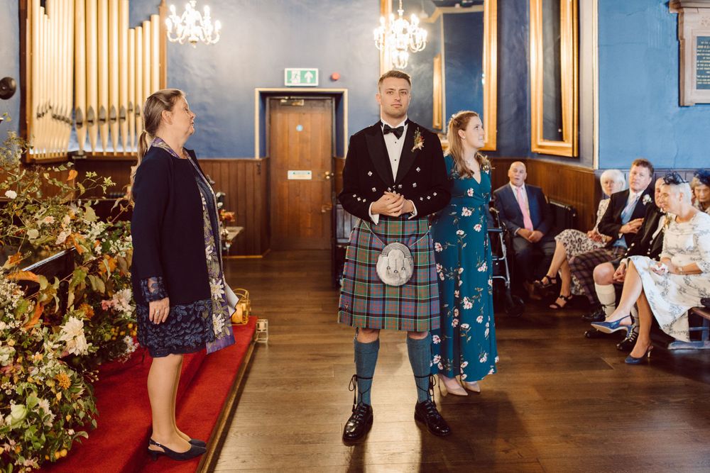 Wedding ceremony in blue-walled room with Scottish kilts and formal attire.