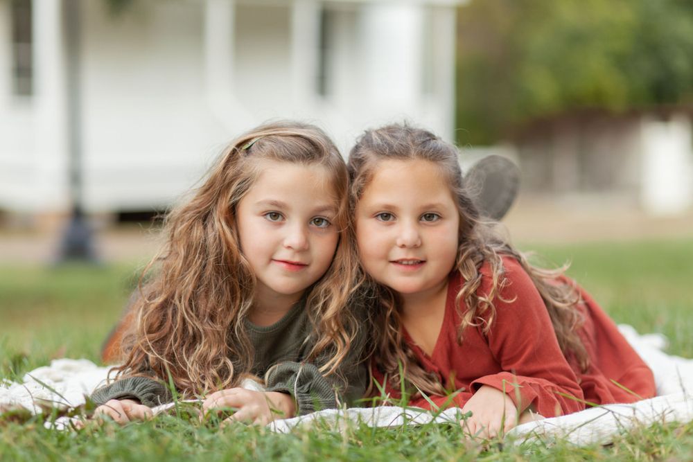 Two young sisters laying on grass together wearing casual clothes and smiling  at Historic Oak View Park in Raleigh.