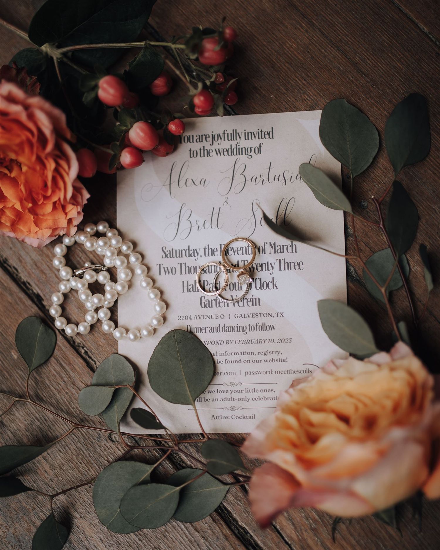 Wedding invitation with rings surrounded by peach roses, eucalyptus leaves, and pearls on wooden surface.