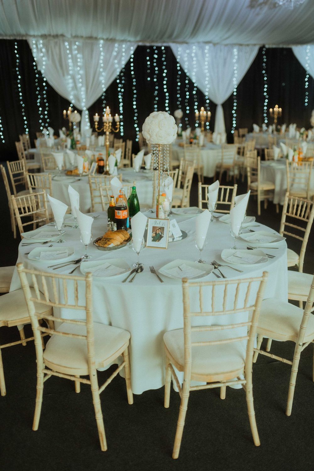 Elegant wedding reception table setting with white linens and chiavari chairs under string lights.