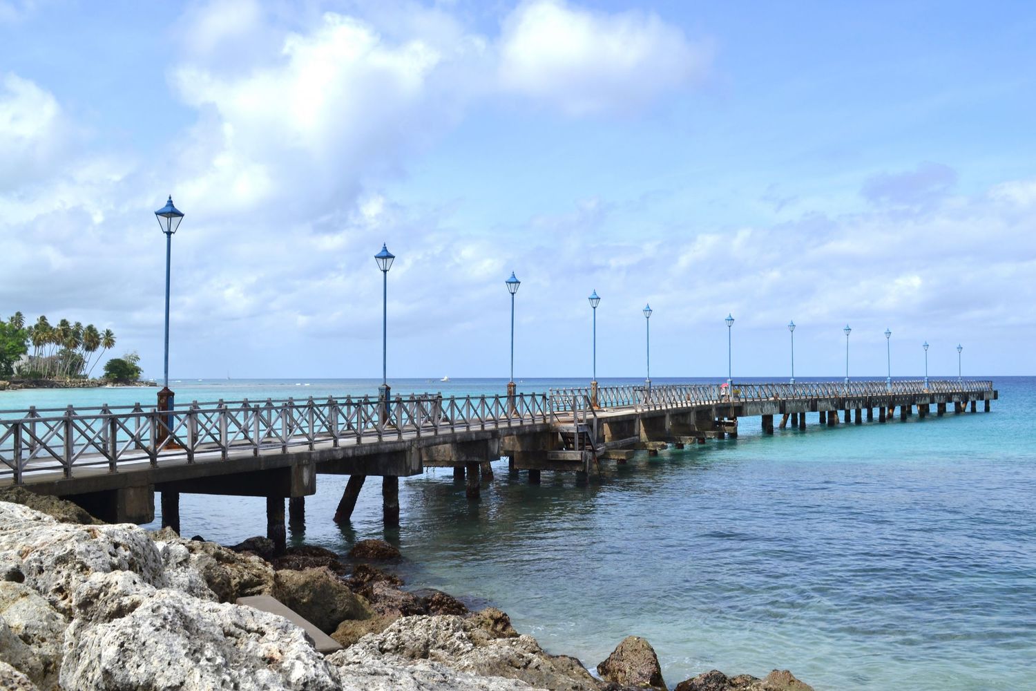 A scenic wooden pier extends over crystal-clear turquoise waters with lamp posts lining the walkway under a partly cloudy sky.
