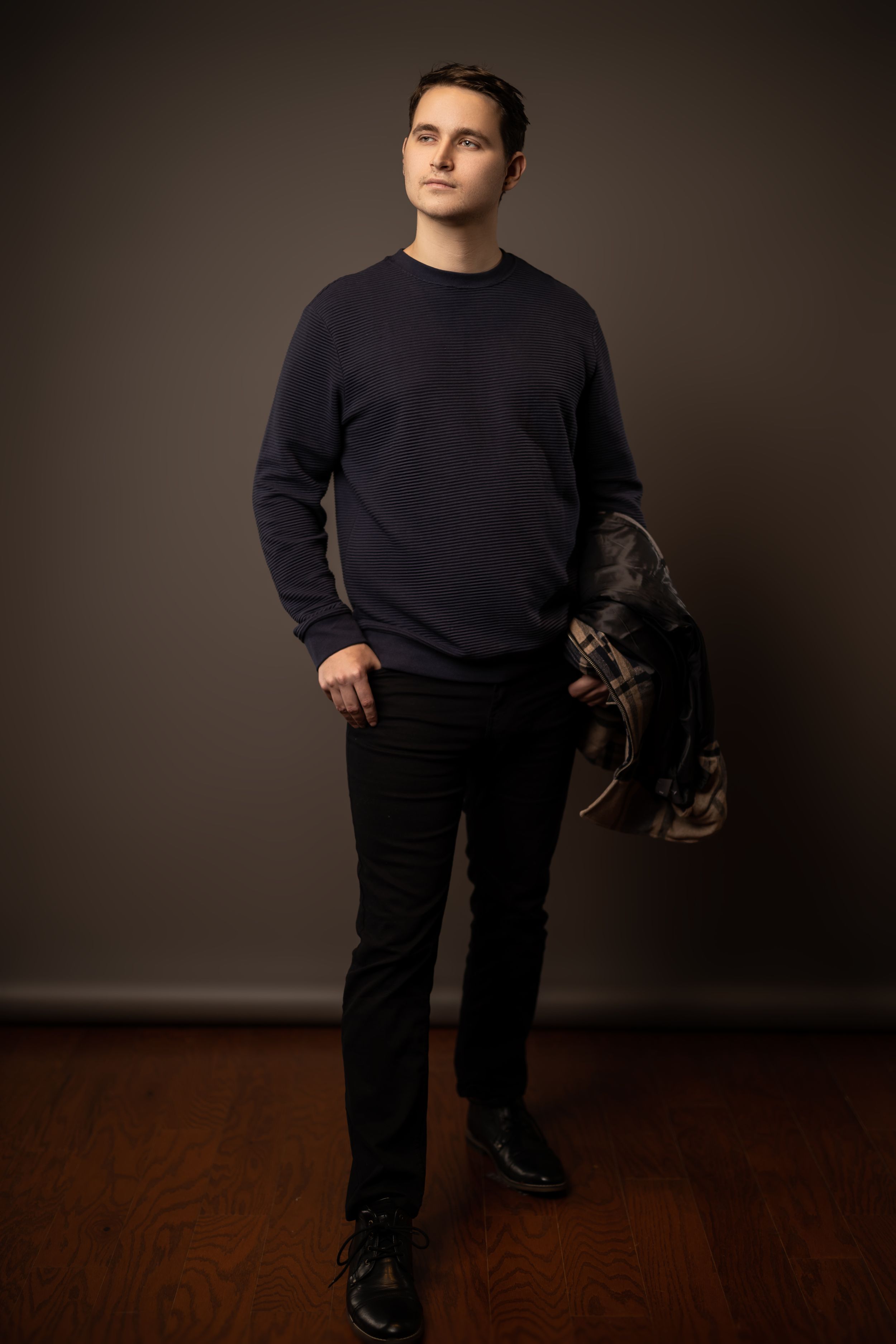 Professional portrait of person in navy sweater and dark pants against brown background.