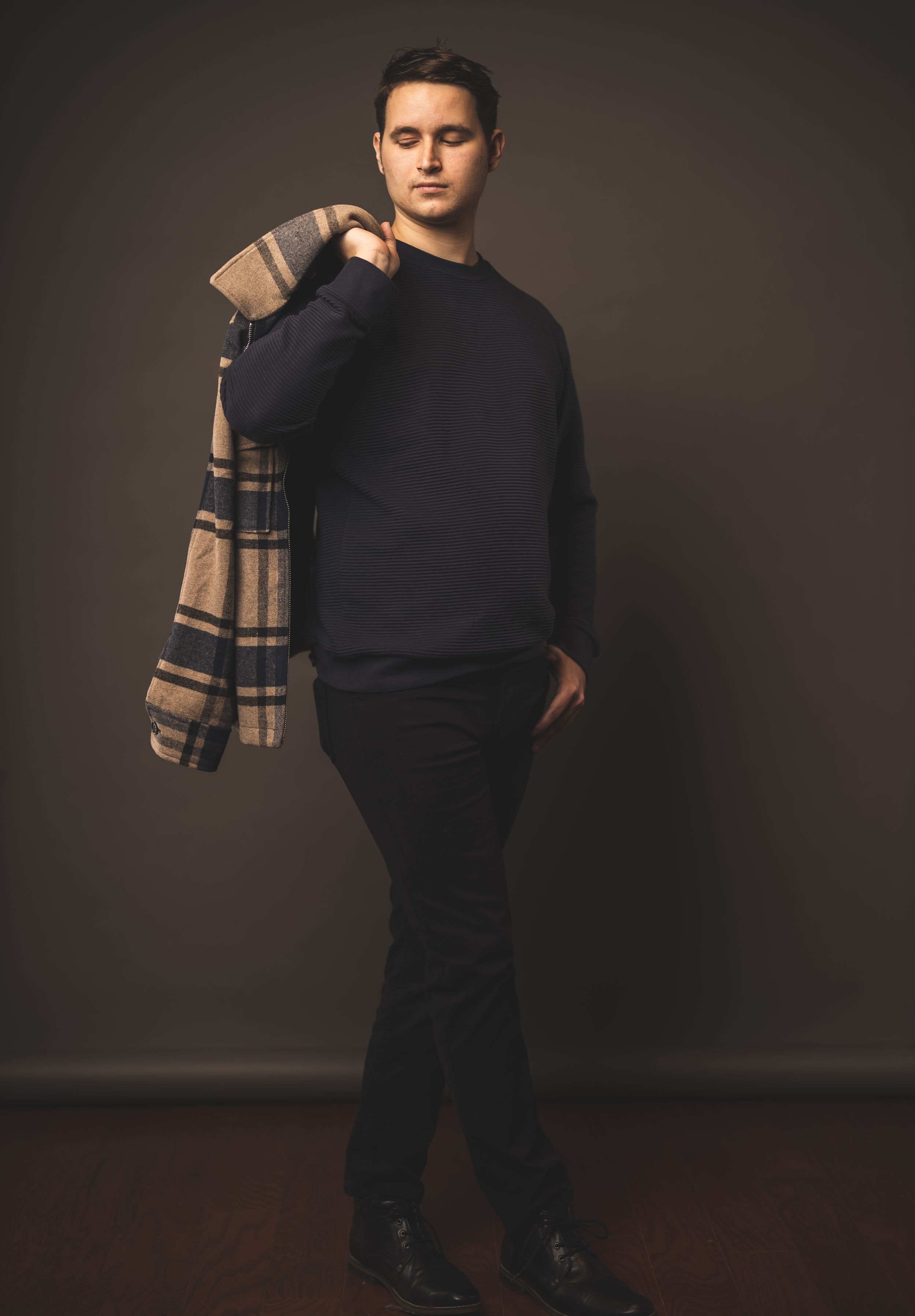 Fashion model wearing dark sweater and beige plaid scarf against brown background in studio portrait.