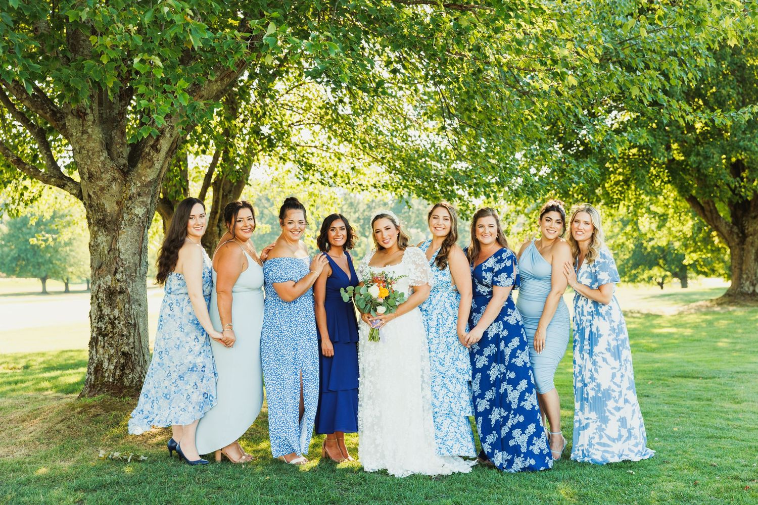 Wedding party members in blue and white dresses gather under green trees for group photos.