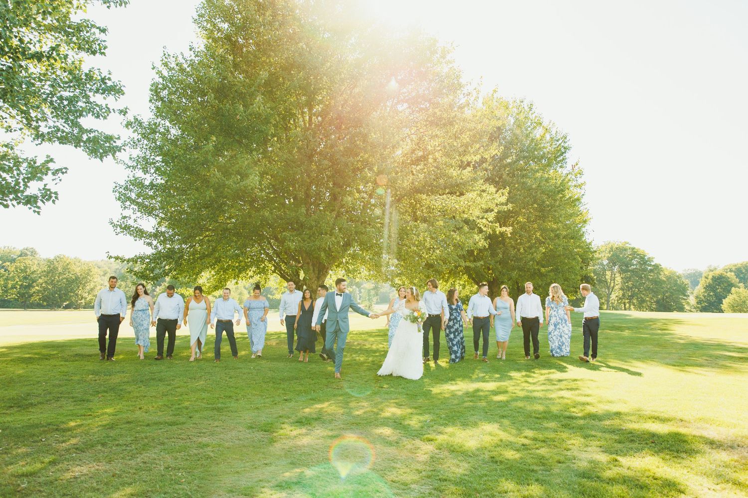 Wedding party gathered on sunlit grassy hill for outdoor group portraits.
