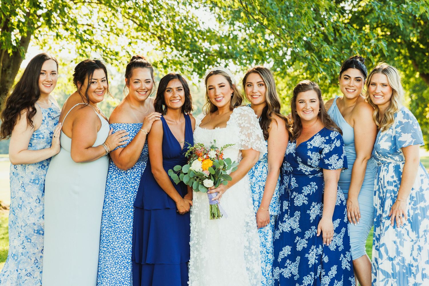 Bridesmaids wearing various blue floral and solid dresses pose together outdoors.