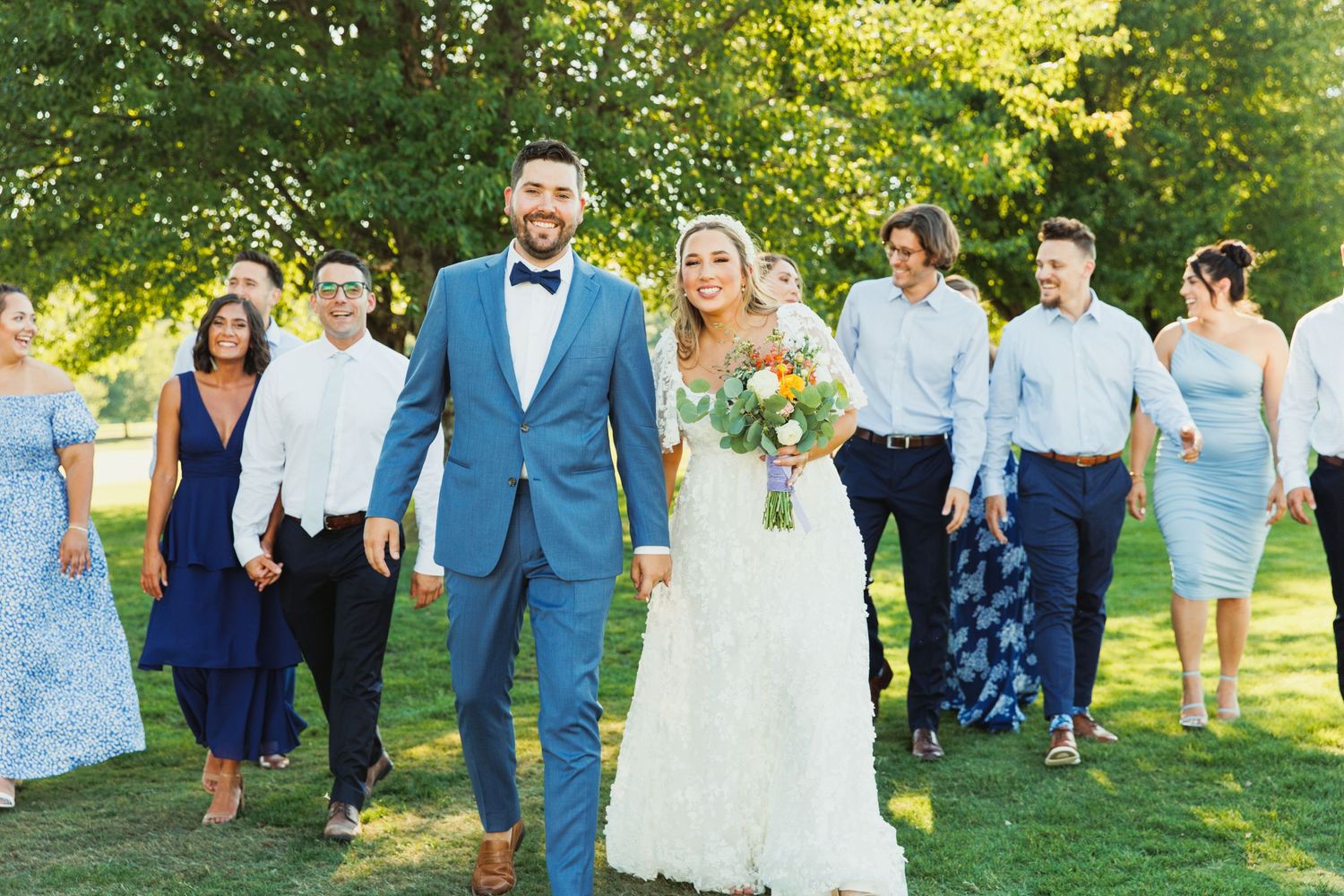 Wedding party in coordinating blue and white attire poses together on a sunny lawn with trees in background.