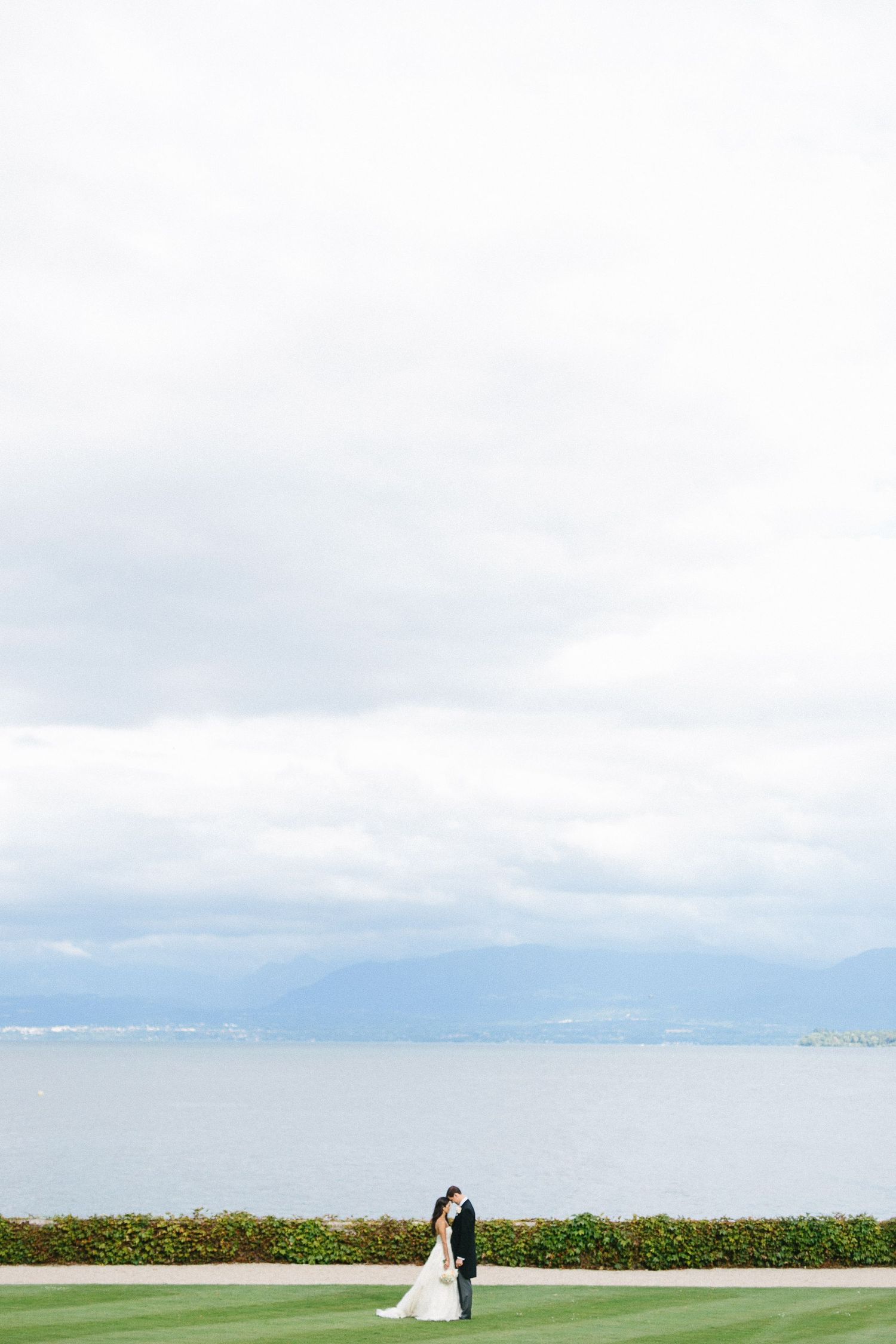 Distant wedding couple on manicured lawn overlooking serene ocean and mountains.