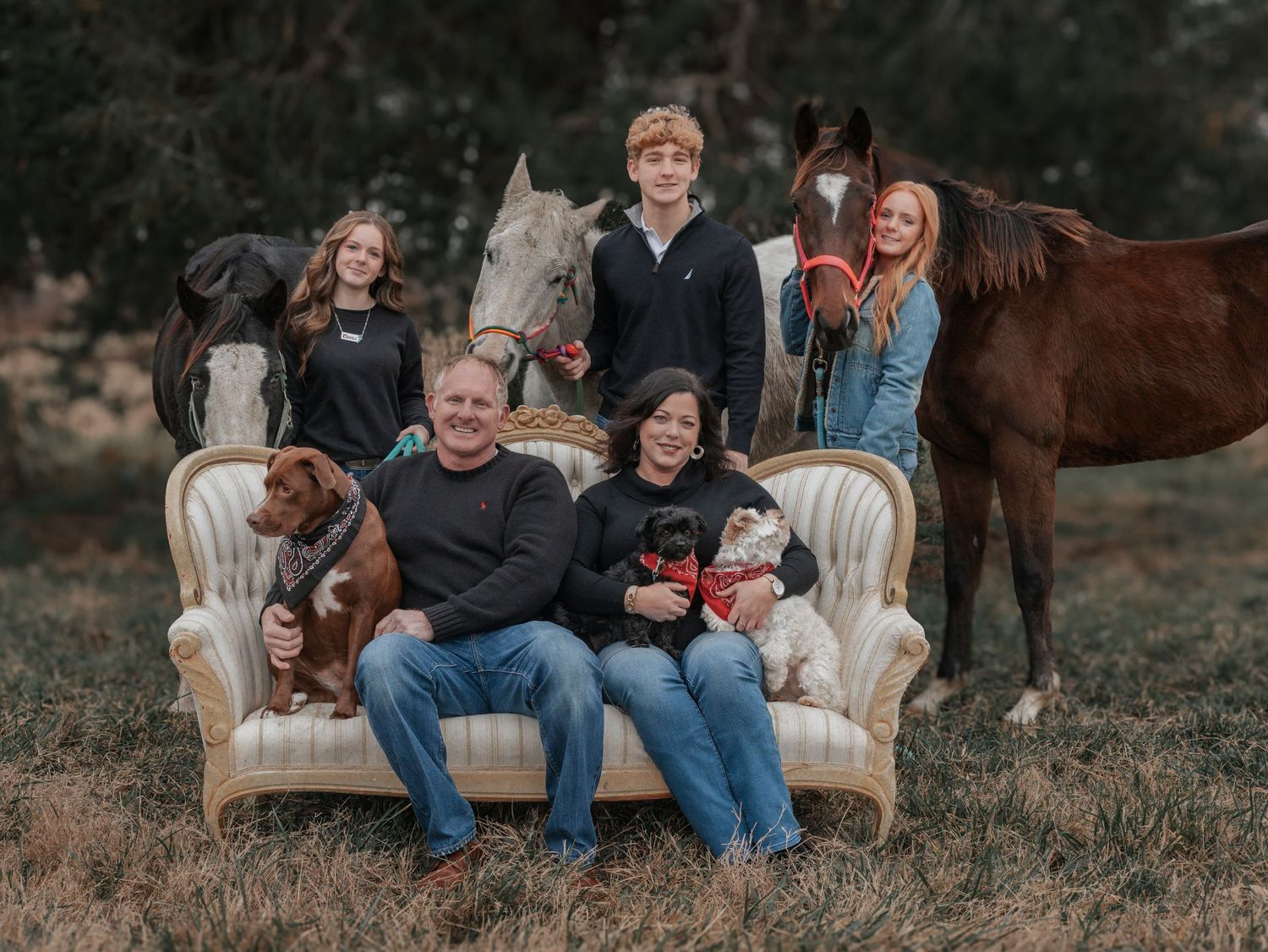 Family posing on vintage couch outdoors with horses in rustic setting.
