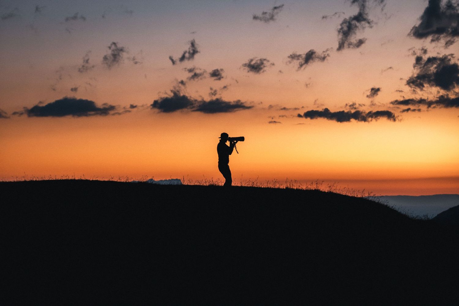 Silhouette of photographer against vibrant orange sunset sky on hilltop.