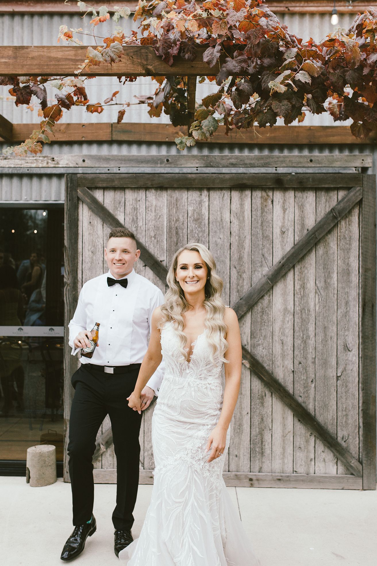 A bride in a fitted lace gown and groom in a tuxedo pose in front of a rustic barn door.