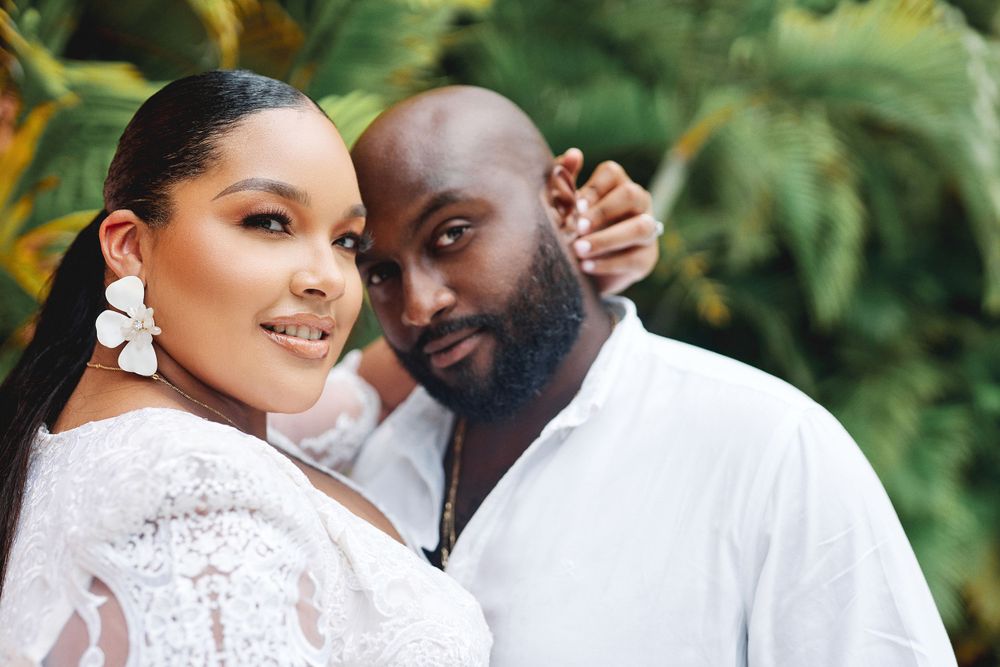 A couple poses together against a tropical backdrop, wearing white outfits in a natural setting.