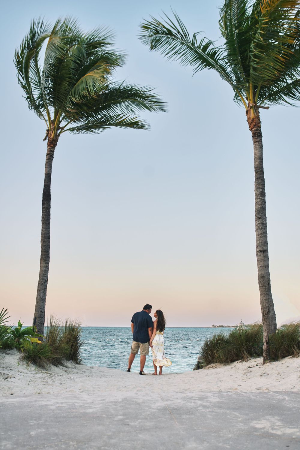 Couple silhouetted between palm trees on white sand beach at sunset.