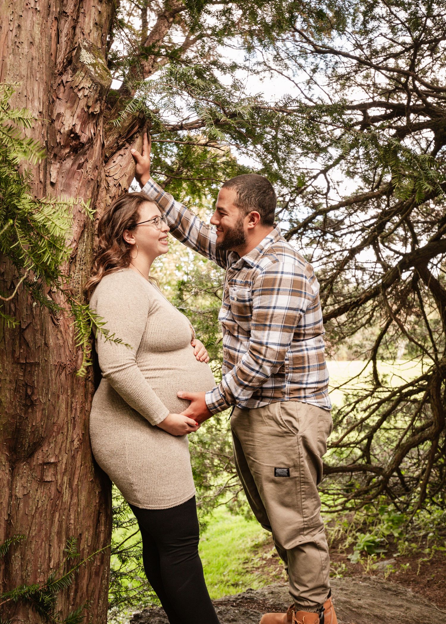 A pregnant couple shares a tender moment leaning against a large tree in a scenic outdoor setting.