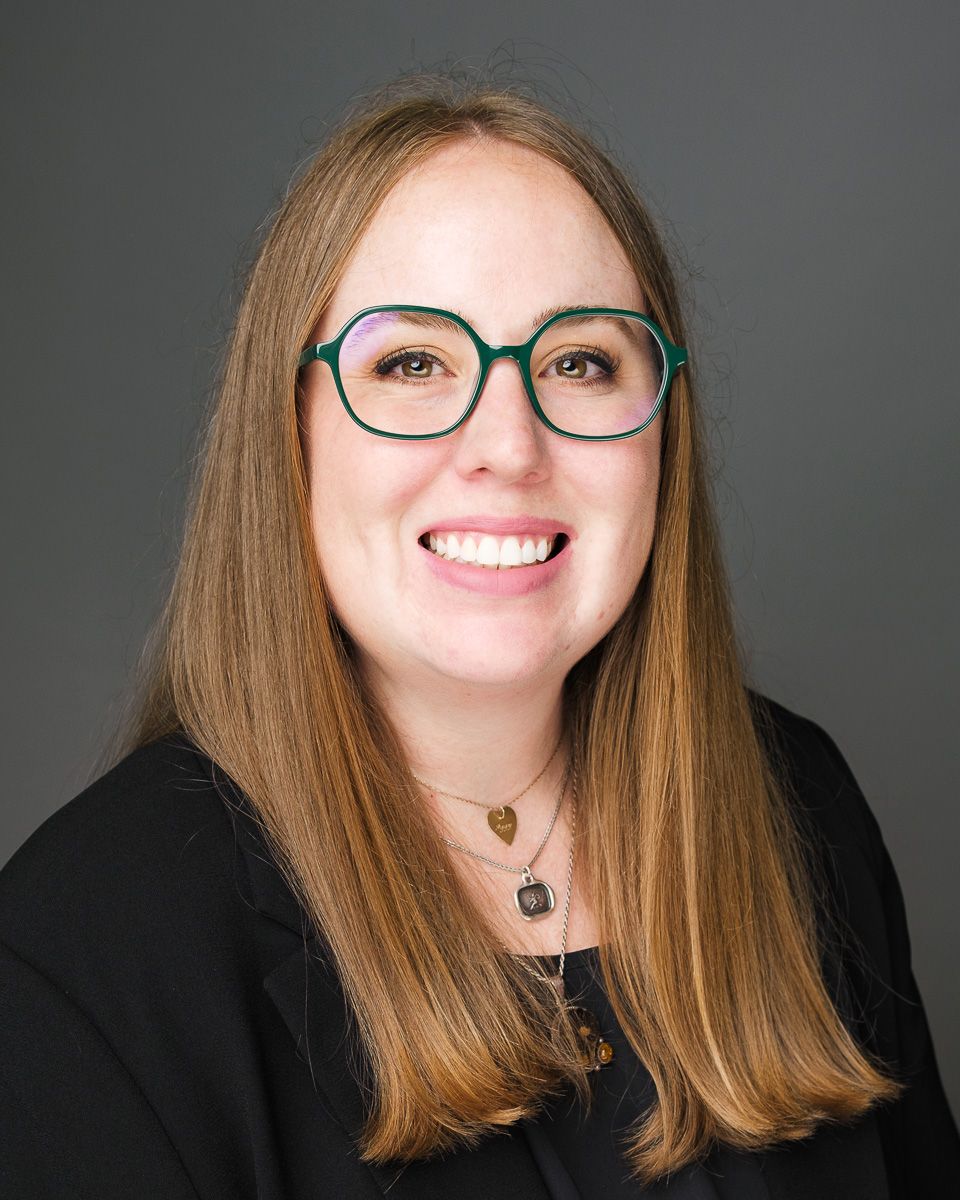 Professional headshot of someone wearing glasses and a black top, smiling warmly against a gray background.
