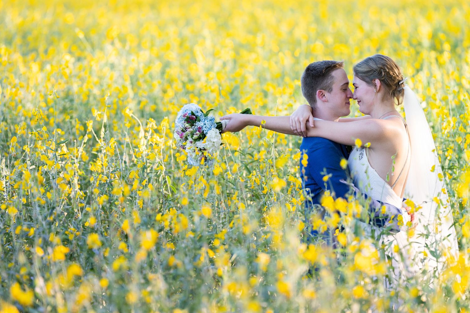 A couple embraces in a field of bright yellow wildflowers holding a bouquet.