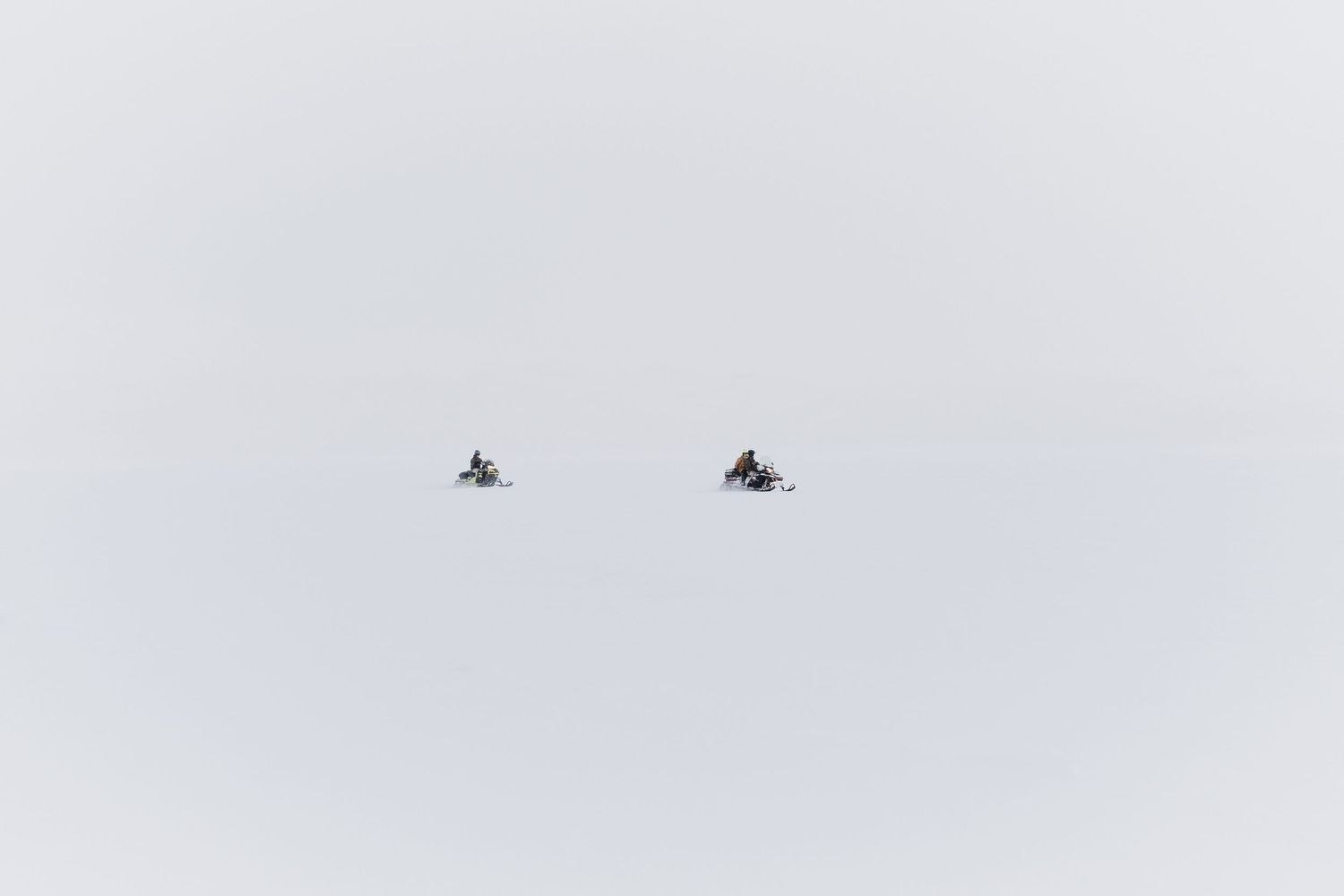 Two snowmobiles traverse across a vast snowy landscape in the distance.