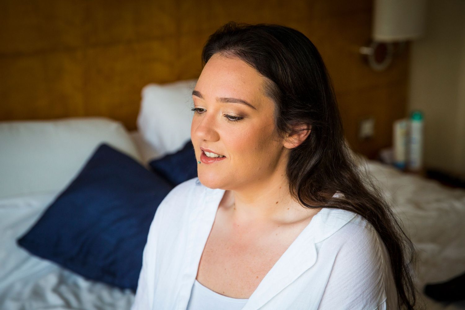Bride in white robe sits by window during wedding day preparations.